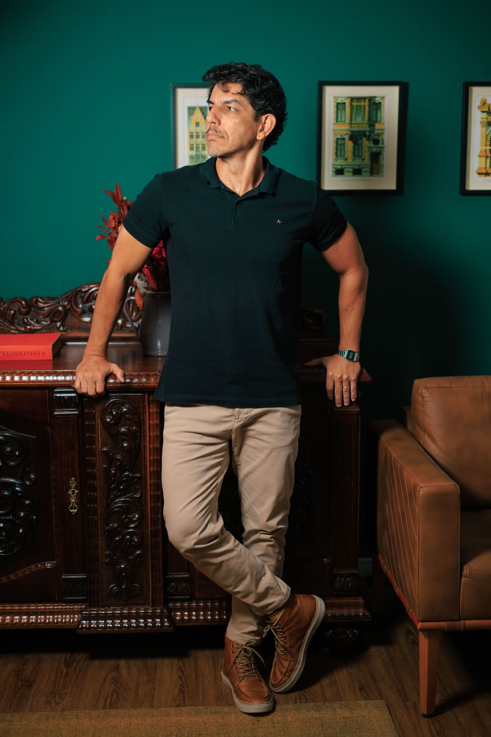 Fred Dias de pé, apoiado em um móvel de madeira entalhada, diante de uma parede verde-garrafa com quadros.