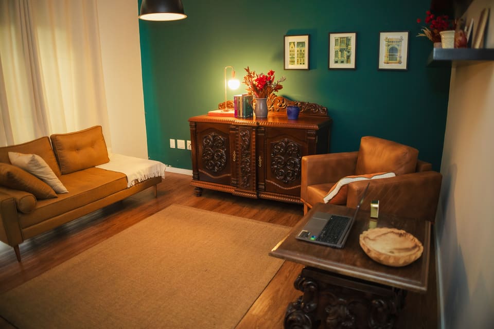 Consultório com parede verde-garrafa, poltrona de couro, sofá caramelo, móvel de madeira entalhada e luminária acesa.