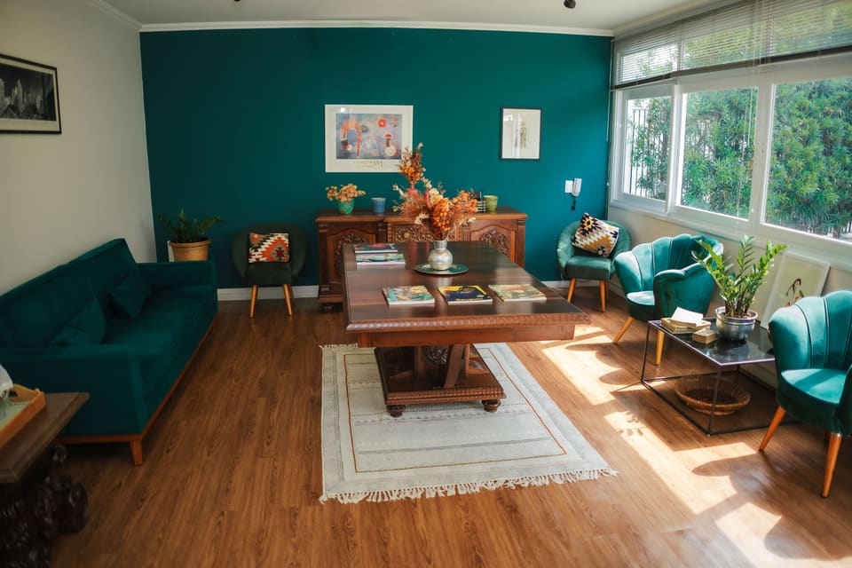 Sala ampla e iluminada, com sofás verdes, mesa de madeira, tapete e janelas para o jardim.